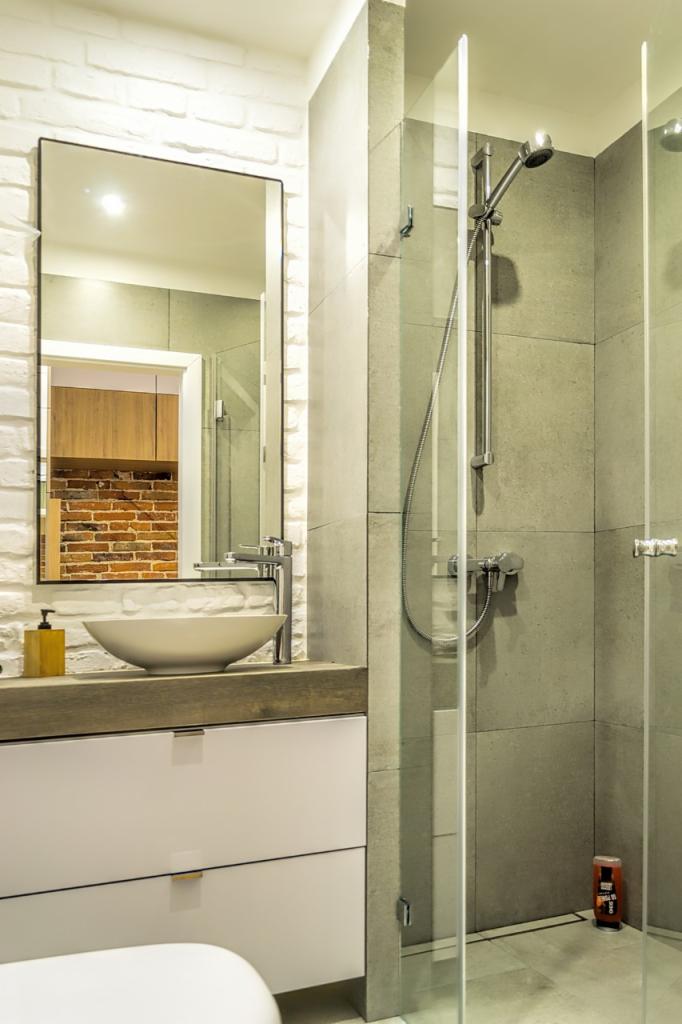 Nowoczesna łazienka z białą cegłą na ścianie, umywalką w kształcie misy na drewnianym blacie i kabiną prysznicową ze szklanymi drzwiami, widok na fragment ściany z czerwonej cegły w lustrze.
