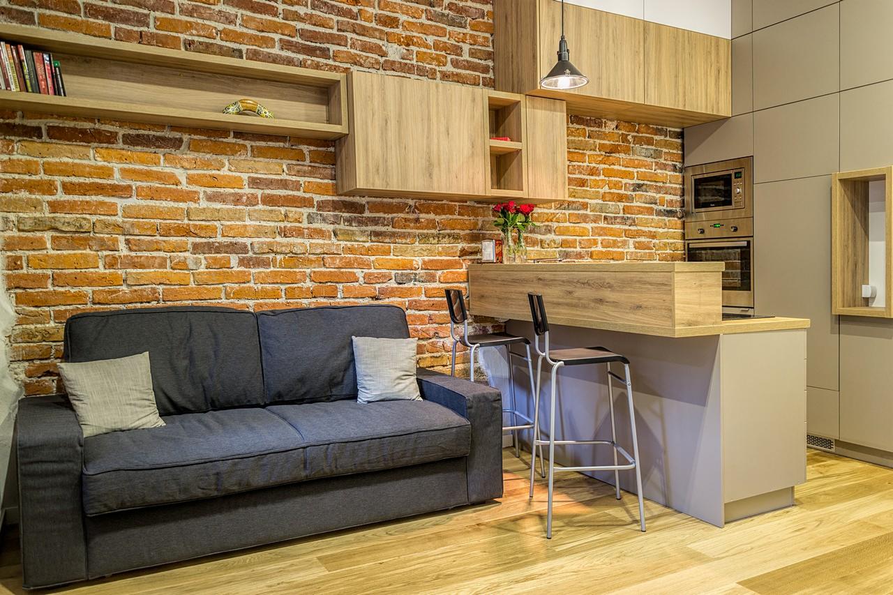 Salon z aneksem kuchennym w stylu loftowym: ceglana ściana, szara sofa, drewniane meble kuchenne i podłoga, nowoczesne oświetlenie.