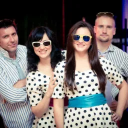 Czteroosobowy zespół w stylizowanych strojach w grochy i okularach pozuje na tle sceny z niebieskim oświetleniem.