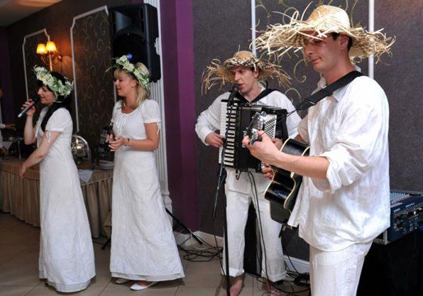 Zespół muzyczny w słomkowych kapeluszach i białych strojach występuje na scenie, wokalistka z mikrofonem, akordeonista i gitarzysta.