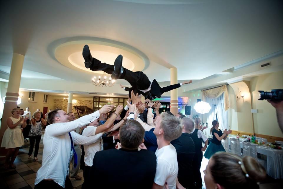 Gość weselny w czarnym garniturze uniesiony przez grupę osób na sali weselnej, w tle widoczny parkiet, oświetlenie i osoby klaszczące.