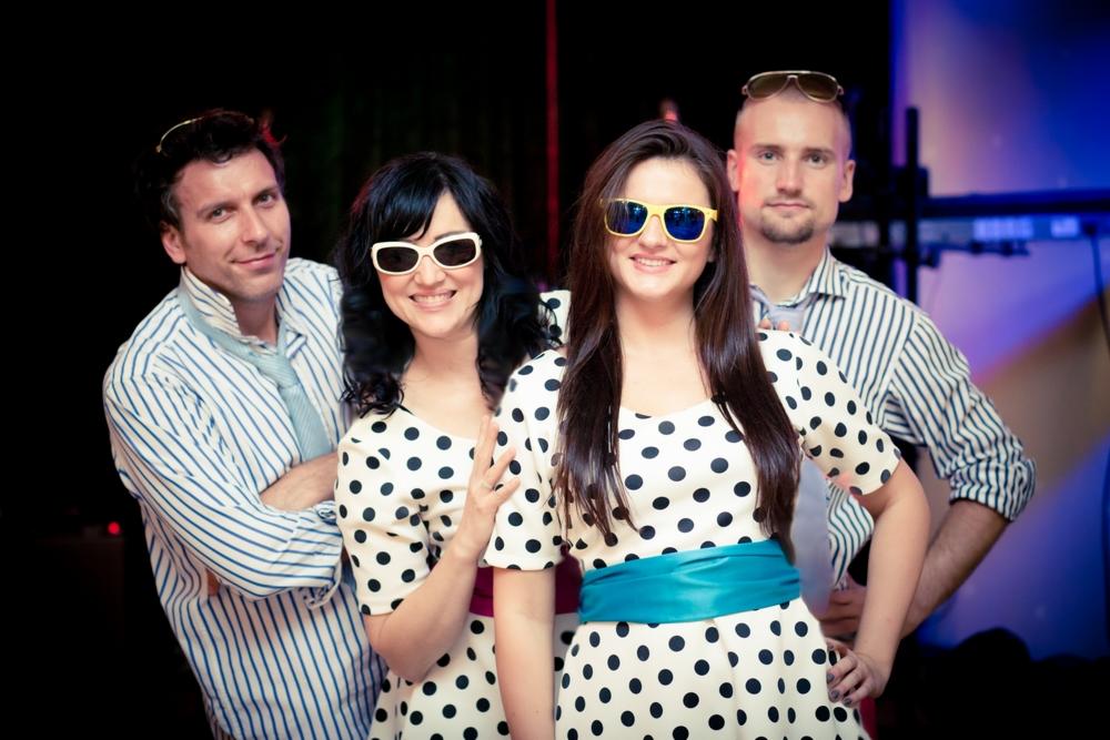 Czteroosobowy zespół w stylizowanych strojach w grochy i okularach pozuje na tle sceny z niebieskim oświetleniem.