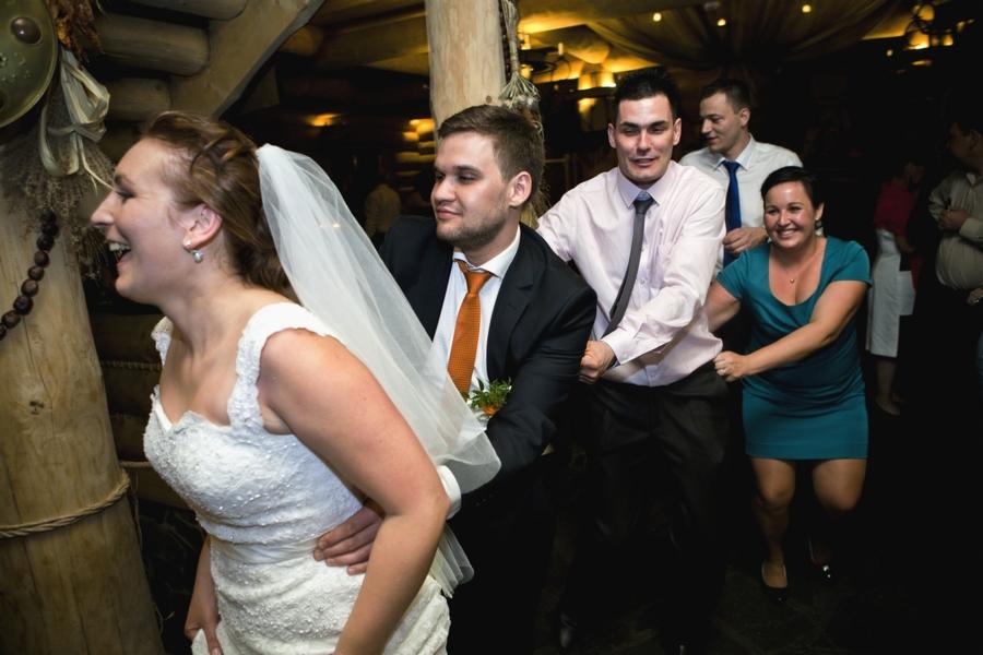 Rozbawiona panna młoda w koronkowej sukni i welonie jest prowadzona w korowodzie przez gości weselnych w trakcie oczepin w sali z drewnianymi elementami wystroju.