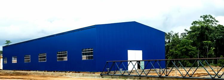 Niebieska hala stalowa z rzędem okien i białymi drzwiami, widoczna z zewnątrz, z metalową konstrukcją pomostu na pierwszym planie i zielenią w tle.