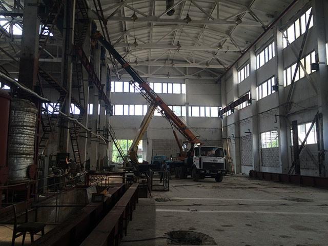 Wnętrze hali przemysłowej z konstrukcją stalową w trakcie montażu przy użyciu dźwigu samochodowego, widoczne elementy instalacji i okna w tle.