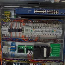 Otwarta szafa sterownicza z widocznymi elementami instalacji elektrycznej, w tym bezpieczniki, styczniki, moduły sterujące i kable różnego koloru, oraz router Netgear na górze, całość oświetlona...