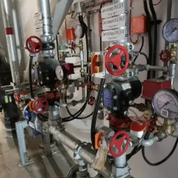 Złożona instalacja hydrauliczna z rurami, zaworami z czerwonymi pokrętłami, manometrami, przewodami elektrycznymi i tabliczkami informacyjnymi w pomieszczeniu technicznym.