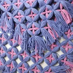 Ręcznie robiona, ażurowa tkanina z niebieskich i różowych włóczek, z dekoracyjnymi frędzlami, ukazująca precyzję wykonania i unikalny wzór.