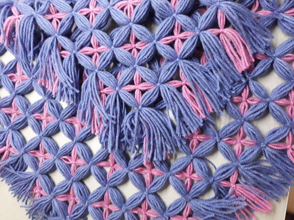 Ręcznie robiona, ażurowa tkanina z niebieskich i różowych włóczek, z dekoracyjnymi frędzlami, ukazująca precyzję wykonania i unikalny wzór.