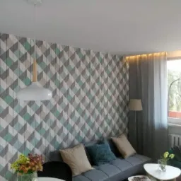 Pokój dzienny z geometryczną tapetą w odcieniach szarości i turkusu, oświetlony nowoczesną lampą wiszącą z drewnianym akcentem, szarą sofą i kwiatami na stoliku kawowym.