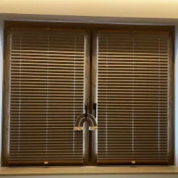 Drewniane okno z brązowymi żaluzjami poziomymi, ozdobione makramowym tęczowym wisiorkiem.