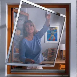 Kobieta montuje moskitierę ramkową w oknie, widoczny pokój dziecięcy z biurkiem i rysunkiem na ścianie.