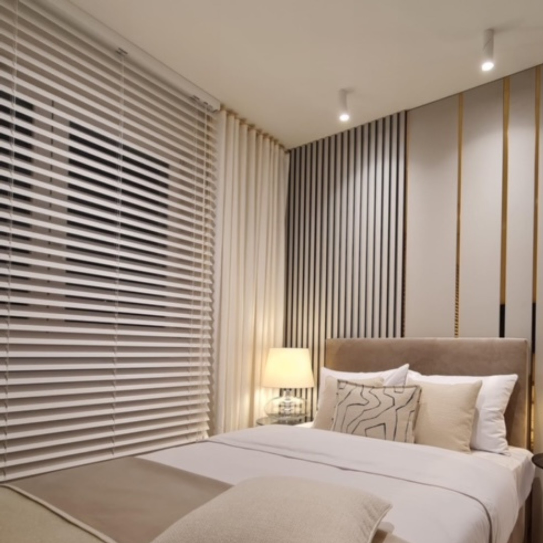 Jasna sypialnia z poziomymi żaluzjami w oknie, beżową pościelą i lampką nocną na szklanym stoliku.