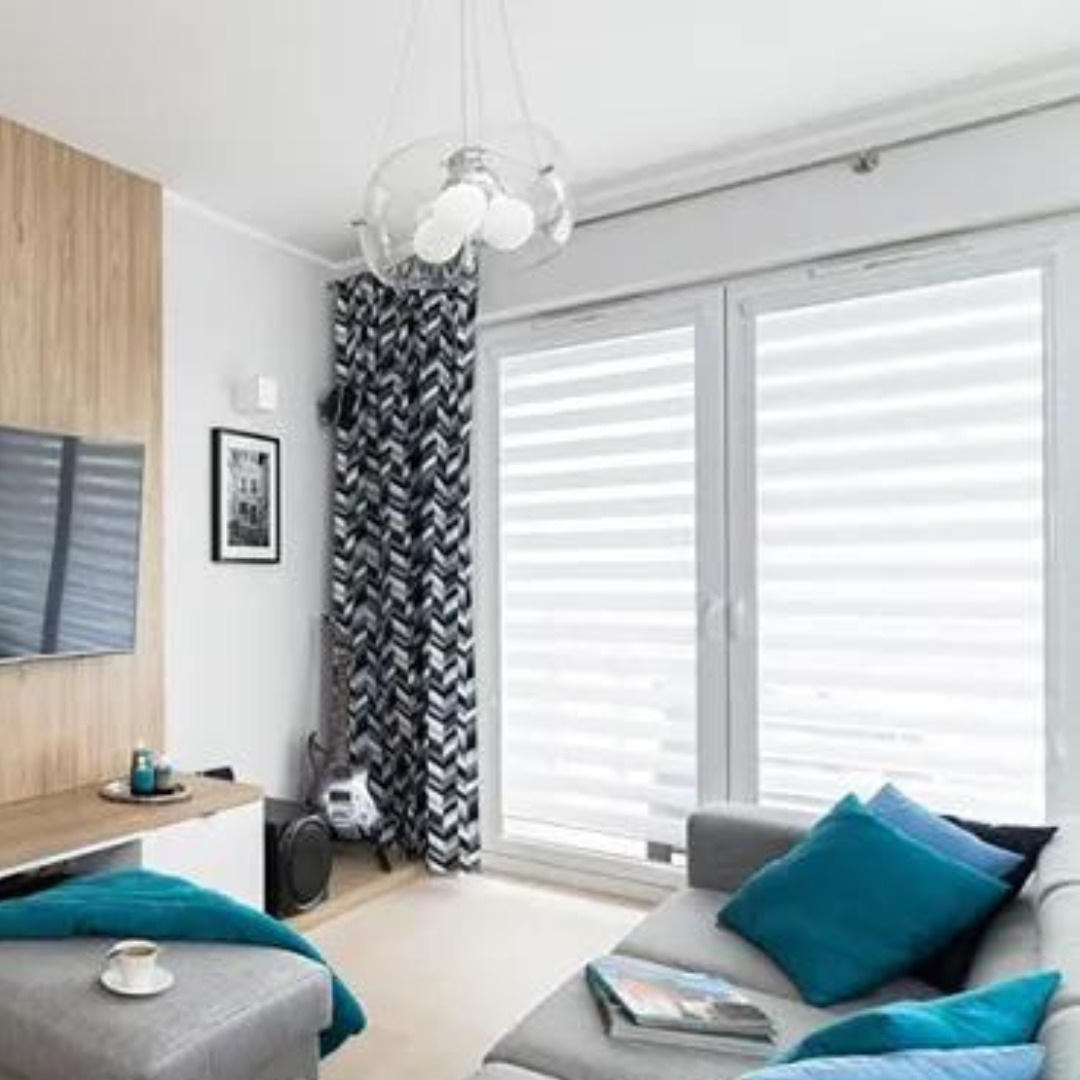 Jasne wnętrze salonu z szarym narożnikiem, turkusowymi poduszkami i kawą na pufie. W oknie podwójne, białe przesłony poziome z delikatnymi paskami, obok zasłona w zygzaki. W tle gitara i lampa...