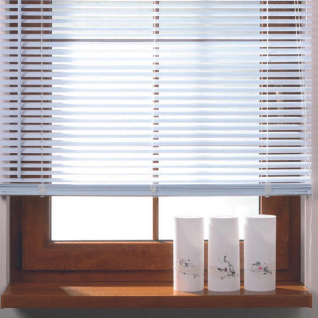 Białe poziome żaluzje w oknie z brązową ramą, trzy białe cylindryczne wazony z delikatnym wzorem na parapecie.