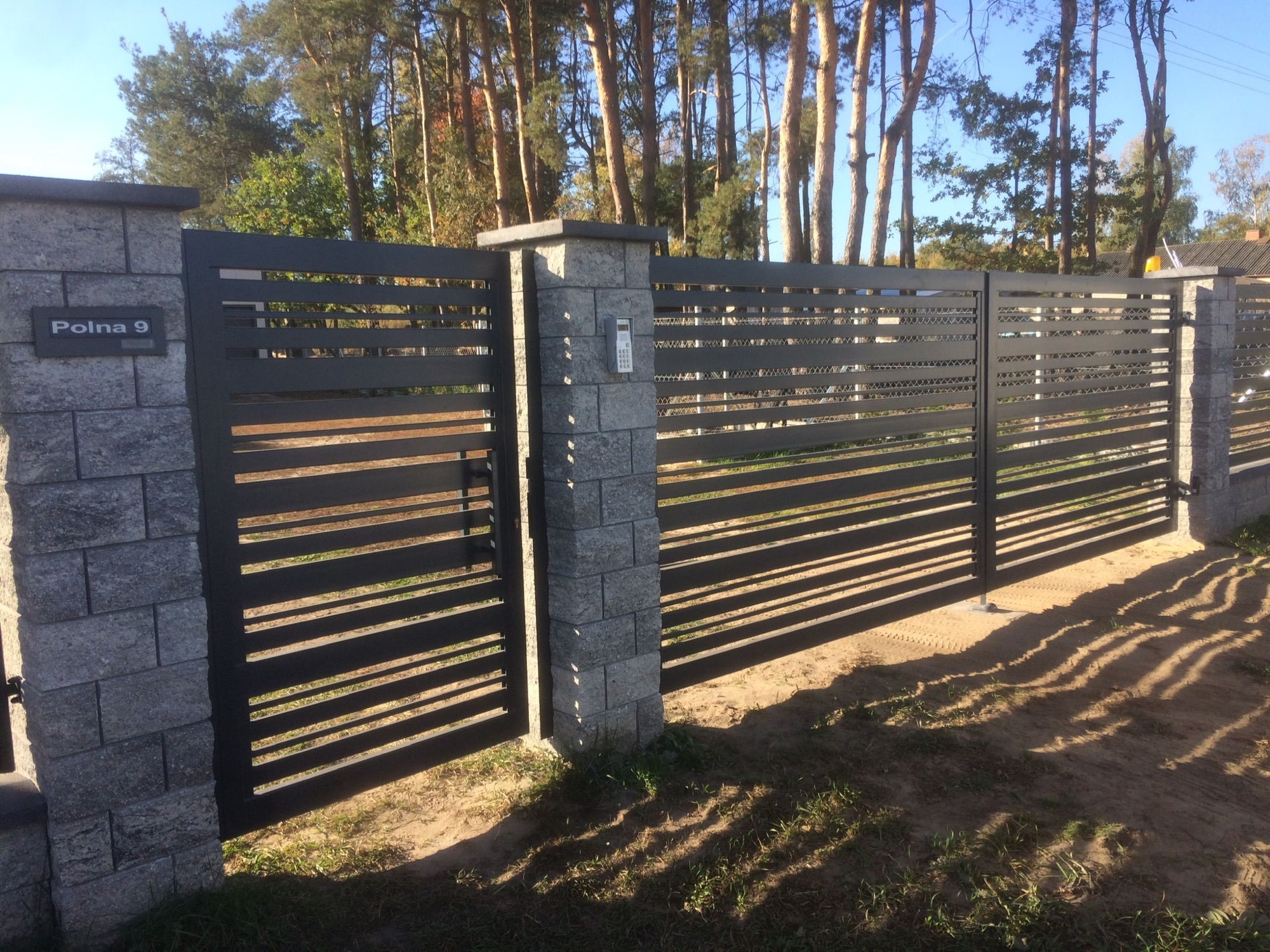 Nowoczesna, pozioma brama wjazdowa i furtka z szarego metalu, zamontowana na słupkach z szarej kostki, tabliczka z numerem 'Polna 9' na słupku, trawa i piasek w tle, drzewa w oddali.