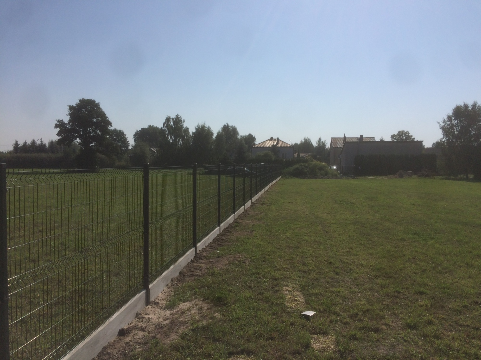 Długi, ciemny, metalowy płot z paneli na betonowej podmurówce oddziela trawnik od posesji z domami w tle, widok z boku.