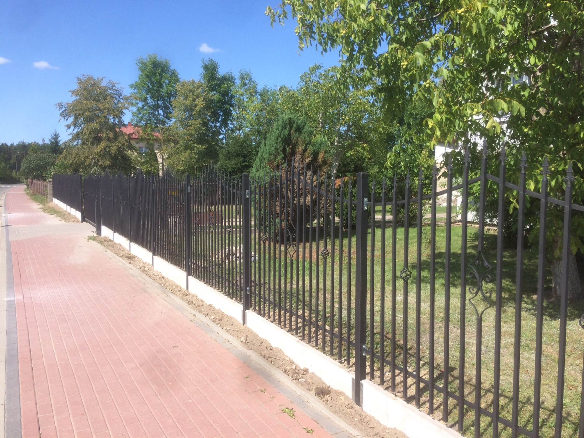 Długie, czarne, metalowe ogrodzenie z ozdobnymi elementami, osadzone na niskiej, betonowej podmurówce, oddziela chodnik z czerwonej kostki od posesji z zielenią i domem w tle.