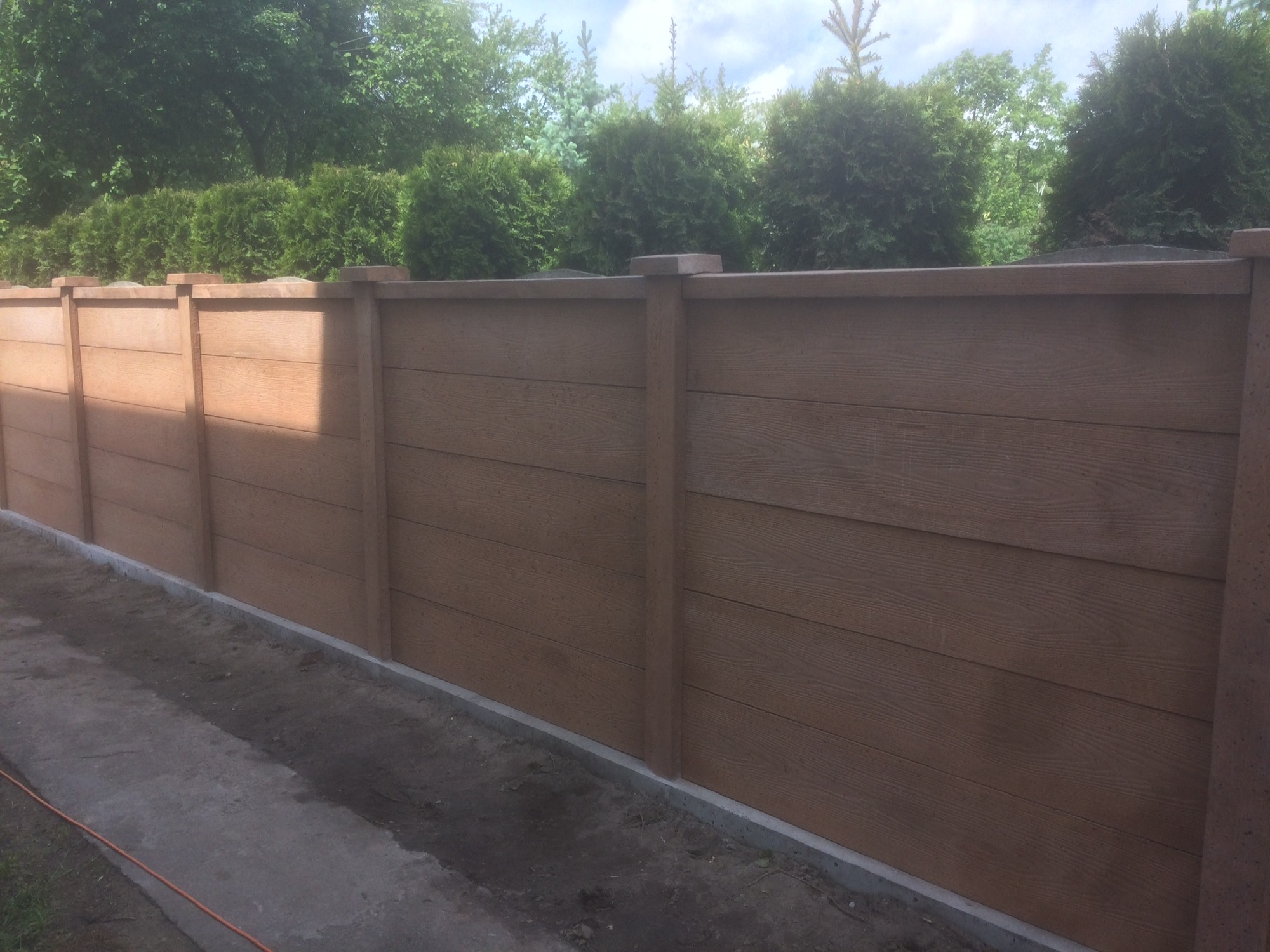 Nowoczesne ogrodzenie z paneli imitujących drewno, widoczne słupki i wypełnienie, w tle zadbane tuje.