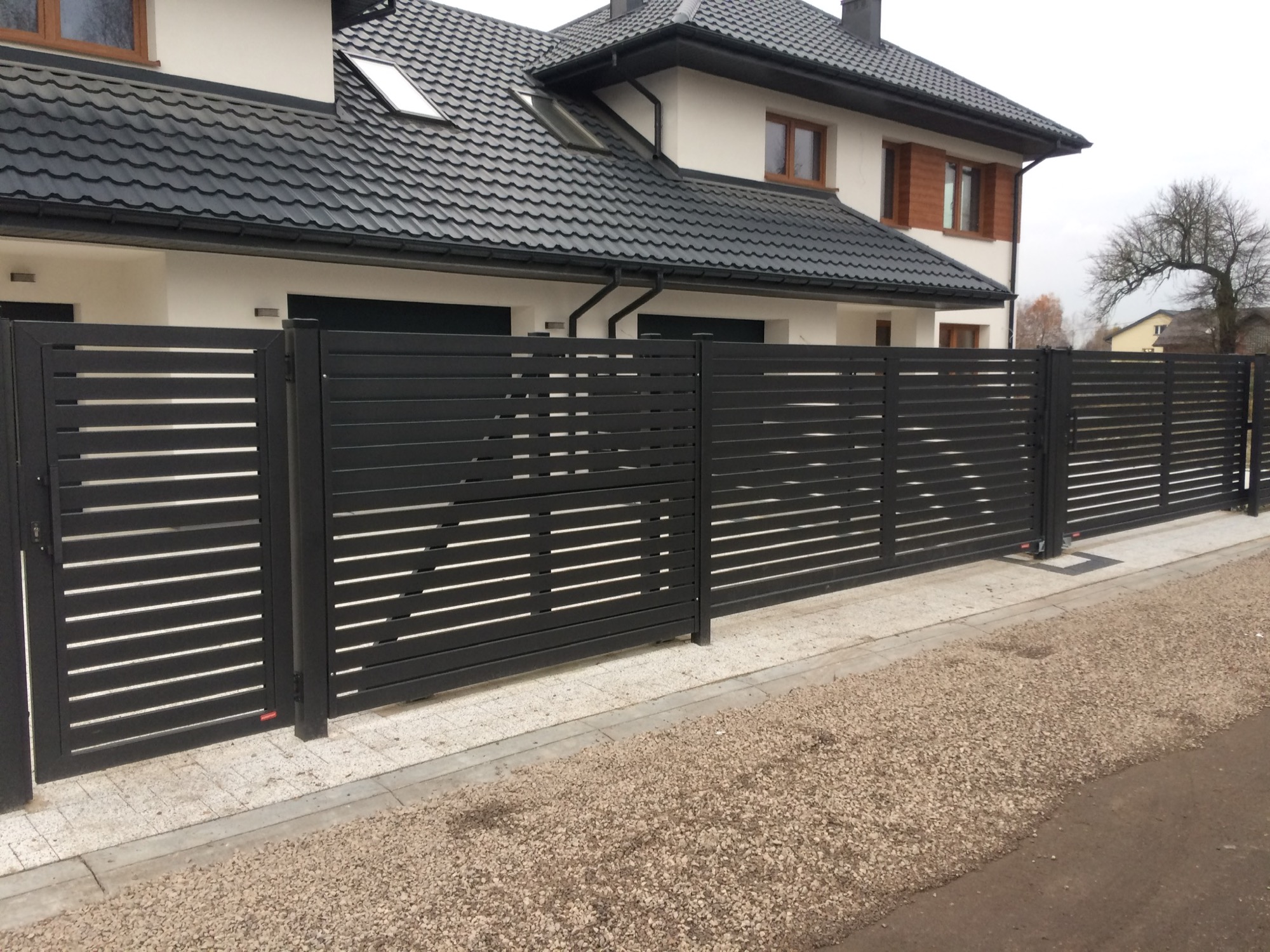 Nowoczesne, poziome, czarne ogrodzenie metalowe z przesuwną bramą wjazdową przed domem z ciemną dachówką i brązowymi ramami okiennymi.