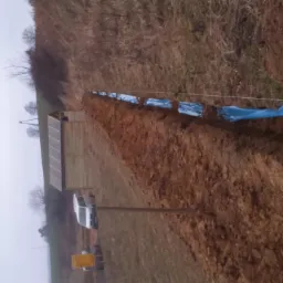 Wykop w ziemi, zabezpieczony niebieską folią i wytyczony sznurkiem, prawdopodobnie pod kable elektryczne, w tle widoczny budynek z panelami słonecznymi.