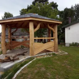 Konstrukcja z surowego drewna w stylu rustykalnym, z dachem krytym papą, na tle budynku gospodarczego i domu jednorodzinnego, fragmenty drewna na posadzce z kostki.