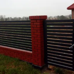 Nowoczesne ogrodzenie panelowe z poziomymi szczeblami w kolorze antracytowym, osadzone na murku z czerwonej cegły, z bramą wjazdową i widocznym fragmentem domu w tle.