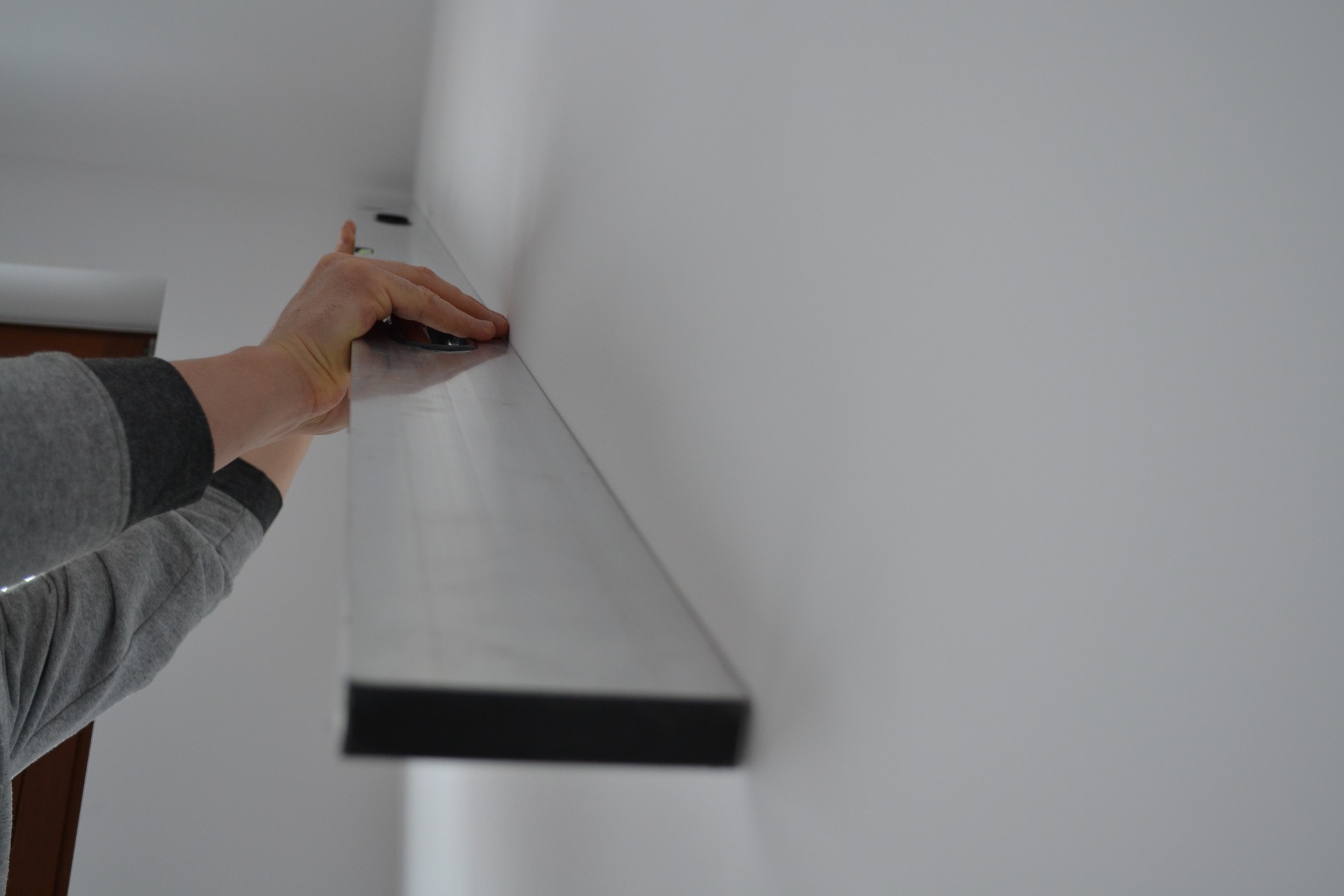 Mężczyzna w szarej bluzie poziomuje długą, ciemną półkę na białej ścianie za pomocą poziomicy, widoczne dłonie i poziomica.