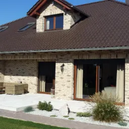 Dom jednorodzinny z jasnej cegły z brązowym dachem, widoczne okna dachowe, rolety zewnętrzne, taras z meblami ogrodowymi i ozdobnym ogrodem.