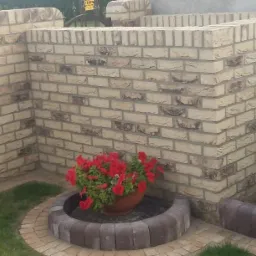 Nowo wybudowany, niski mur z jasnej cegły klinkierowej, otaczający rabatę z czerwonymi kwiatami, wykończony obrzeżem z kostki.