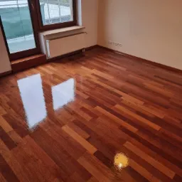 Lśniąca, świeżo polakierowana parkietowa podłoga w pokoju z oknem balkonowym i grzejnikiem, widoczne odbicie światła w lakierze.