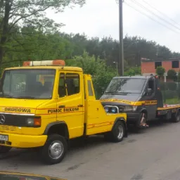 Żółta laweta Mercedes-Benz z napisem 'POMOC DROGOWA' holuje szaro-czarnego busa w zabudowie lawety na tle zieleni i niedokończonego budynku.