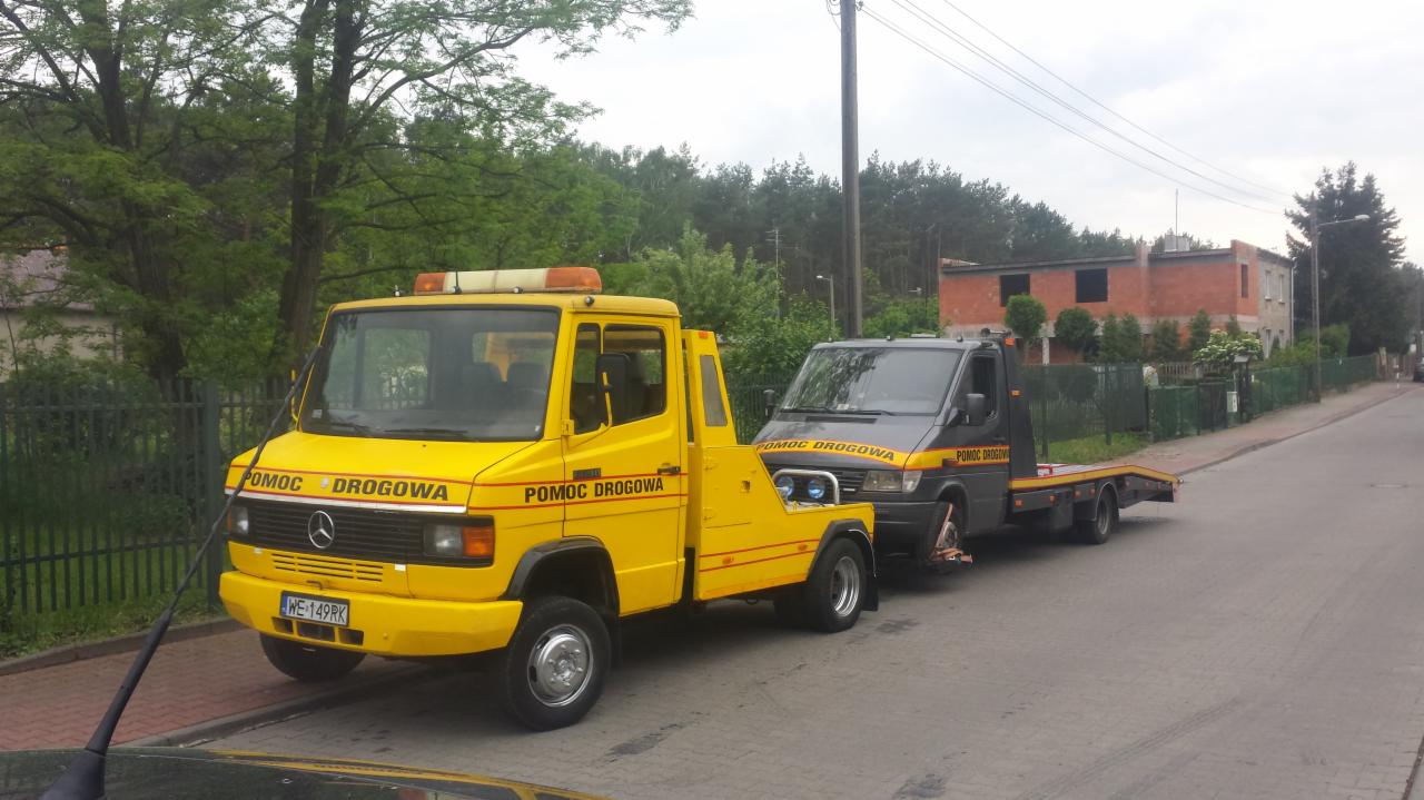 Żółta laweta Mercedes-Benz z napisem 'POMOC DROGOWA' holuje szaro-czarnego busa w zabudowie lawety na tle zieleni i niedokończonego budynku.