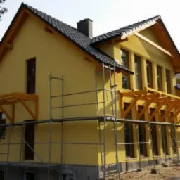 Żółty dom jednorodzinny w trakcie budowy, z widocznymi rusztowaniami i elementami konstrukcji dachu. Dach pokryty ciemną dachówką ceramiczną, okna w trakcie montażu. Elewacja w jasnym odcieniu żółci.