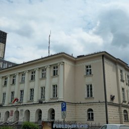 MiM Income Sp. z o.o. - Elewacja budynku Urzędu Dzielnicy Śródmieście w Warszawie, widok z dołu, z flagą Polski i znakami drogowymi. Na tle pochmurne niebo i nowoczesny budynek.