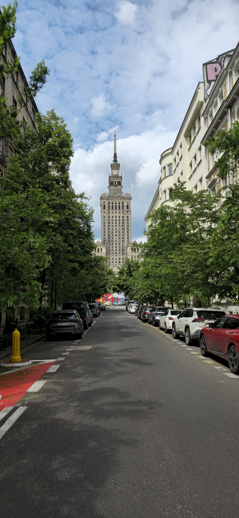Ulica z zaparkowanymi samochodami prowadząca do Pałacu Kultury i Nauki w Warszawie, widzianego pomiędzy drzewami w słoneczny dzień.