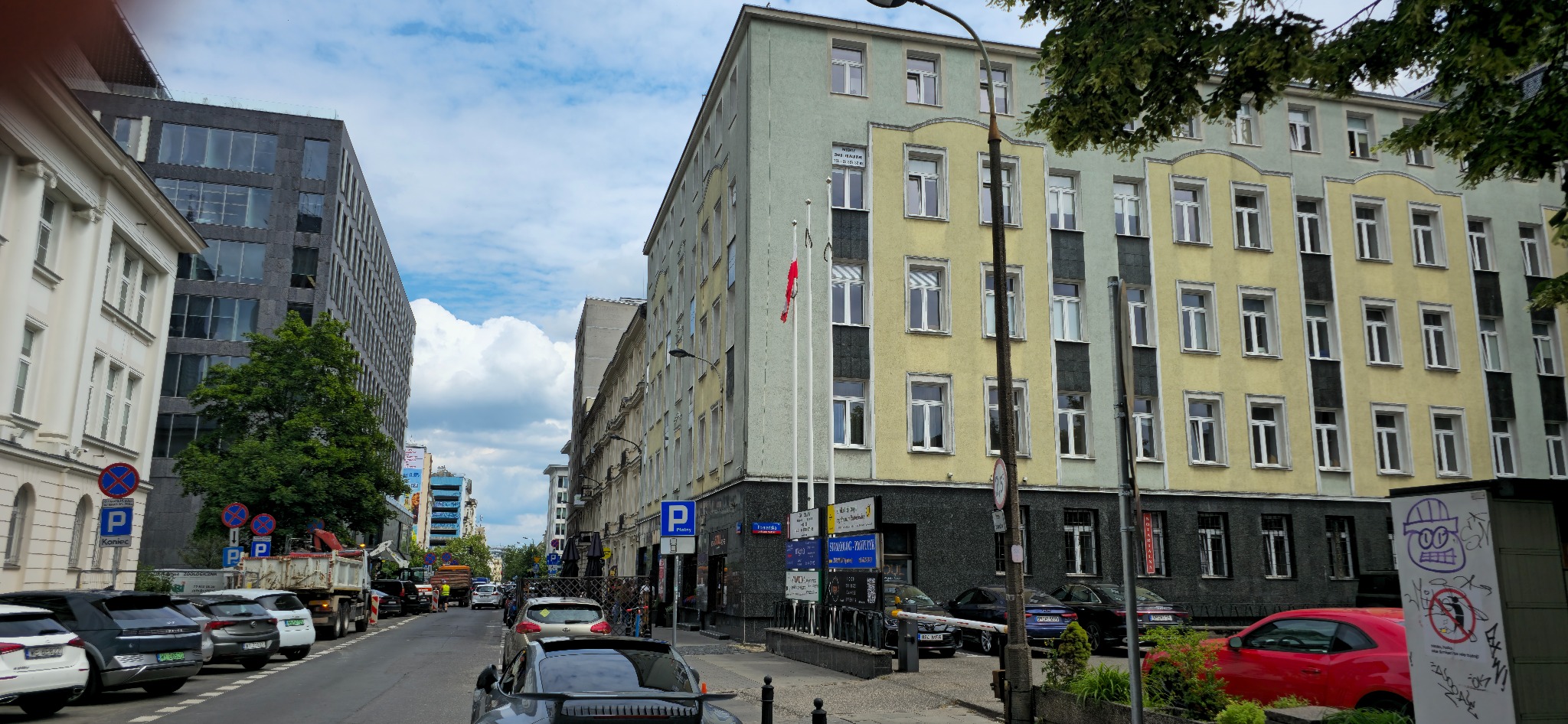 Ulica miejska z budynkami o zróżnicowanej architekturze, zaparkowanymi samochodami i znakami drogowymi w słoneczny dzień. Widoczne flagi i roślinność.