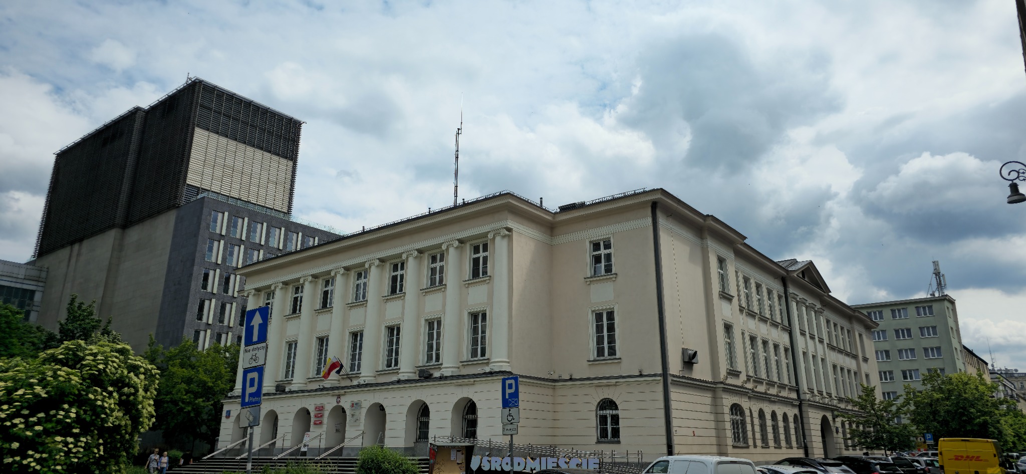 Elewacja budynku Urzędu Dzielnicy Śródmieście w Warszawie, widok z dołu, z flagą Polski i znakami drogowymi. Na tle pochmurne niebo i nowoczesny budynek.