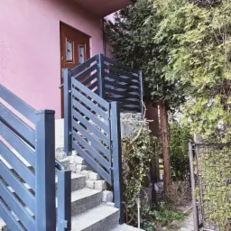 Metalowa balustrada schodowa w kolorze grafitowym, z poziomymi elementami, prowadząca do wejścia domu o różowej elewacji i brązowych drzwiach, otoczona zielenią.