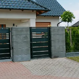 Nowoczesna, pozioma brama wjazdowa z czarnymi, metalowymi przęsłami i betonowymi słupkami, prowadząca do domu z ciemną dachówką i kostką brukową na podjeździe.