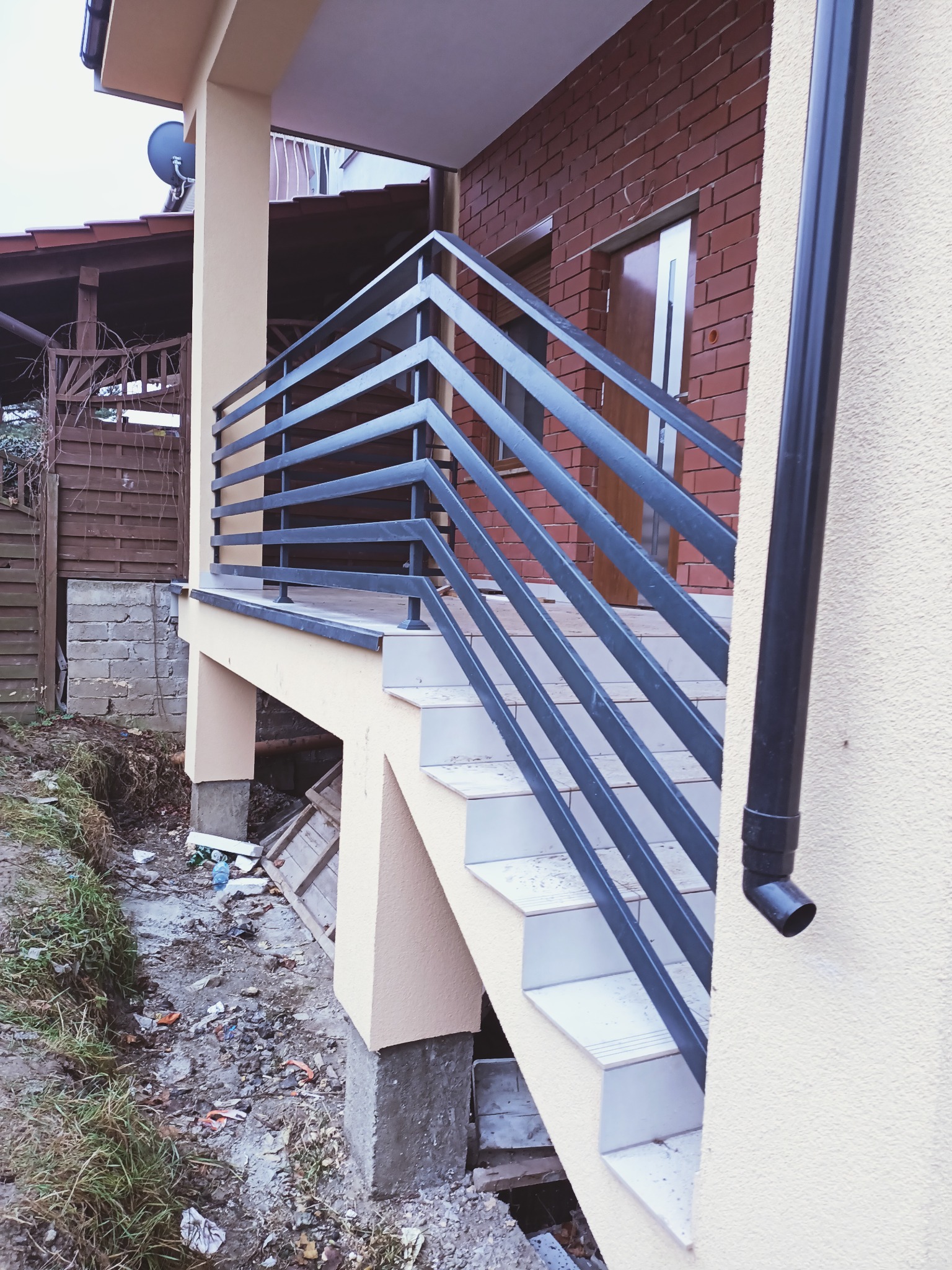 Zewnętrzne schody z metalową balustradą o poziomych prętach, prowadzące do wejścia budynku z ceglaną ścianą i brązowymi drzwiami. Widoczny fragment rynny i teren wokół schodów w trakcie...