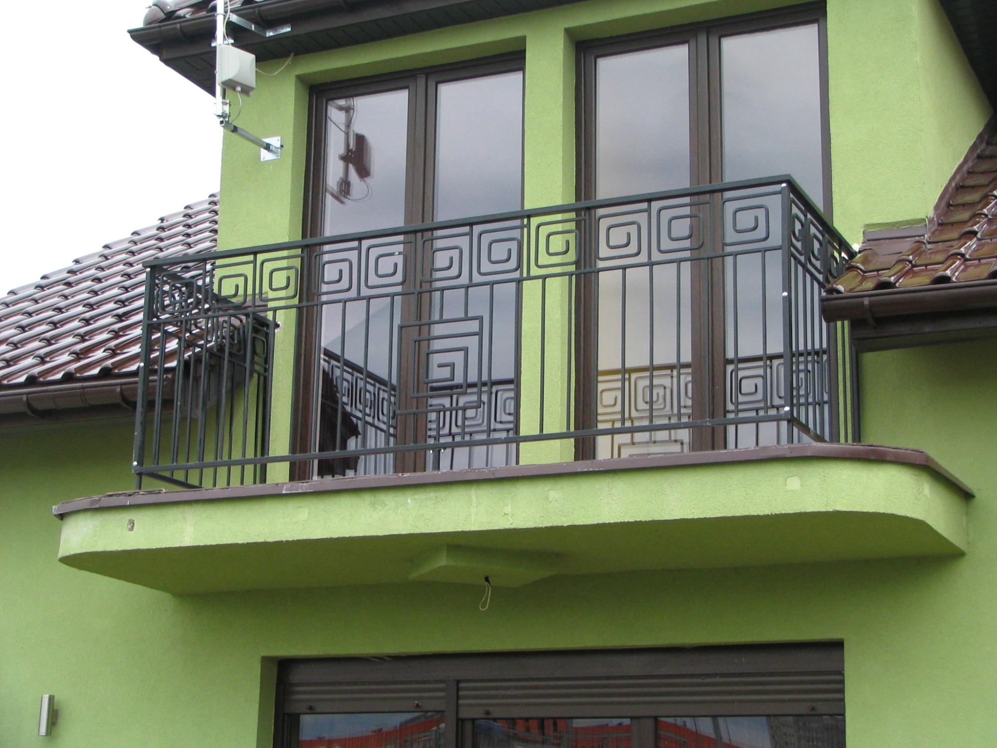 Czarna metalowa balustrada balkonowa z motywem meandru na balkonie budynku w kolorze pistacjowym, widok z dołu.