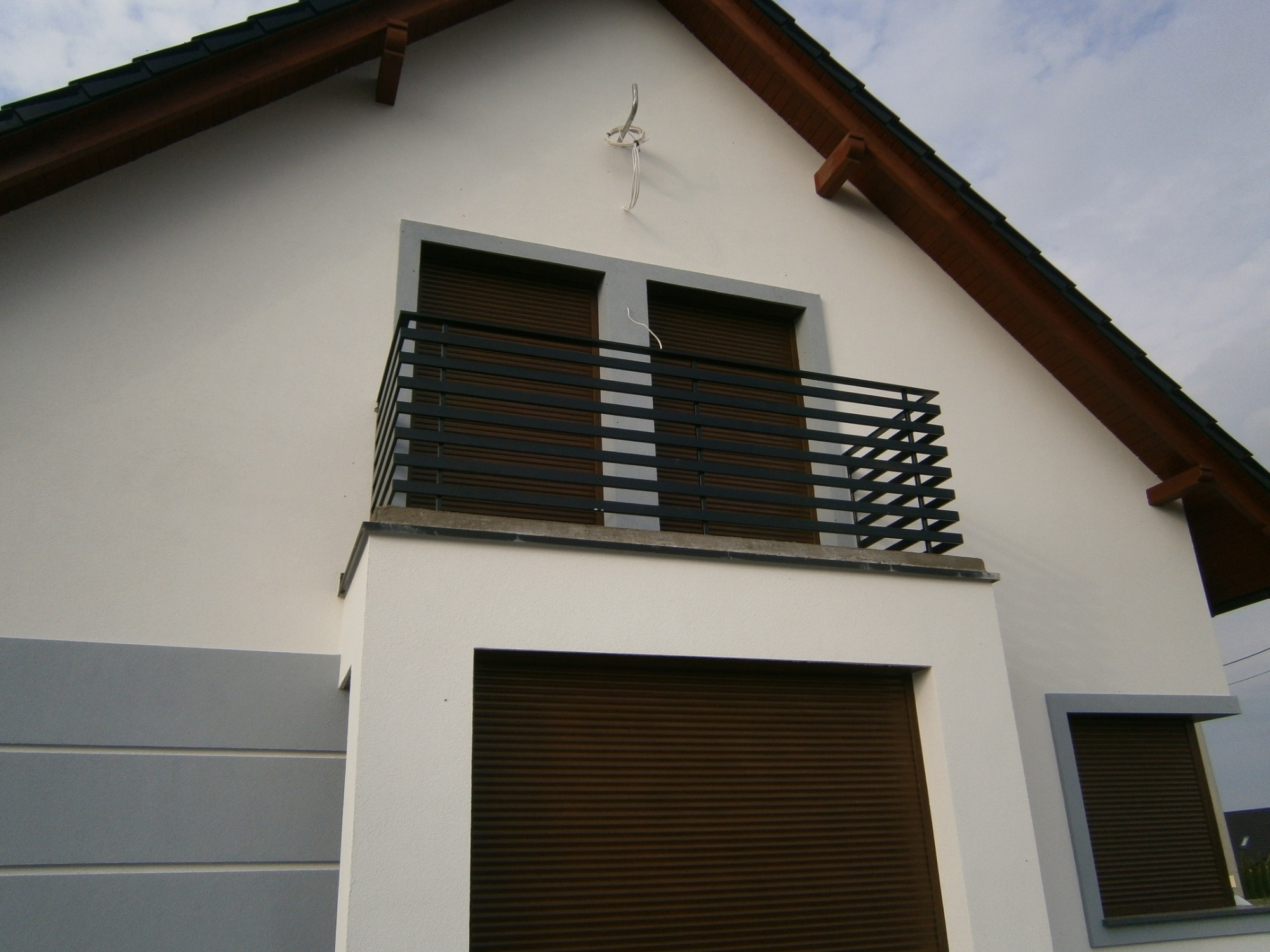 Nowoczesny dom z balkonem zabezpieczonym czarną, metalową balustradą o prostym wzorze, brązowe rolety zewnętrzne na oknach, biała elewacja z szarymi akcentami.