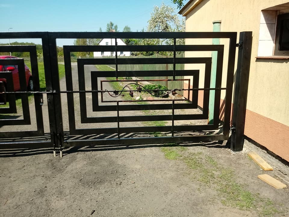 Czarna, metalowa brama wjazdowa o geometrycznym wzorze kwadratów, z ozdobnym elementem roślinnym w centralnej części, na tle domu i zieleni.