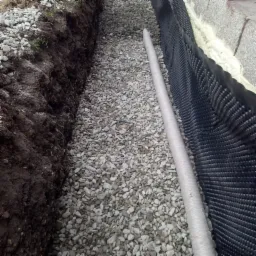 Wykop z rurą drenażową obsypany żwirem, zabezpieczony folią kubełkową przy fundamencie.