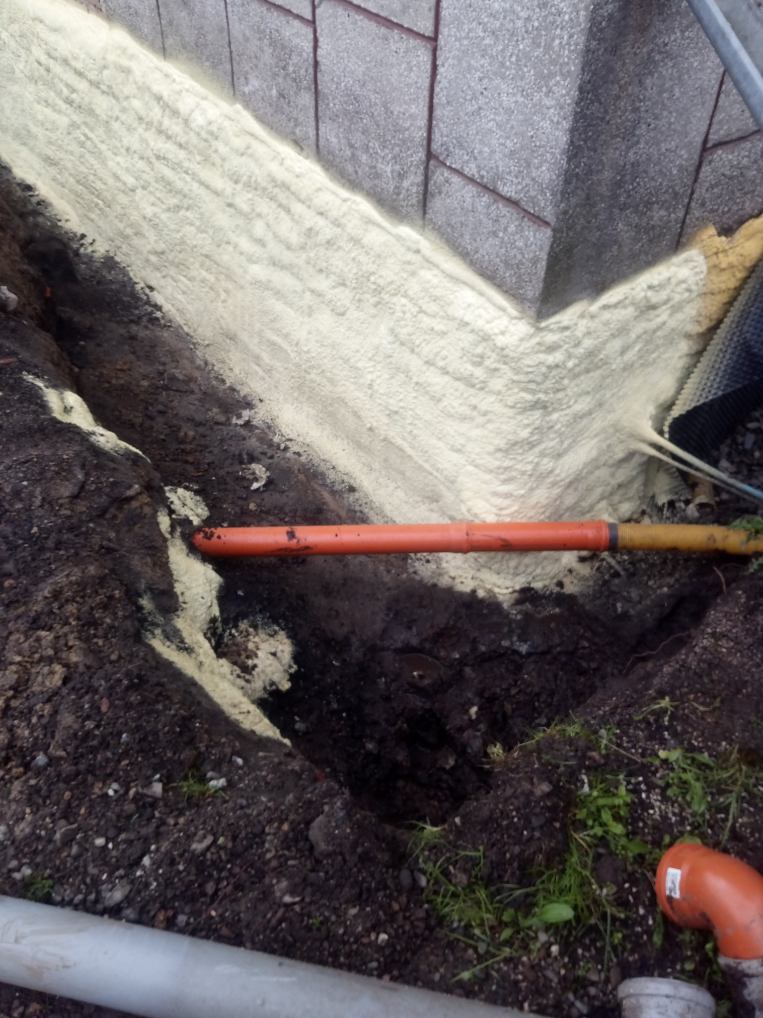 Świeżo nałożona pianka izolacyjna na fundament budynku, częściowo zasypana ziemią, z widocznymi rurami kanalizacyjnymi w wykopie.