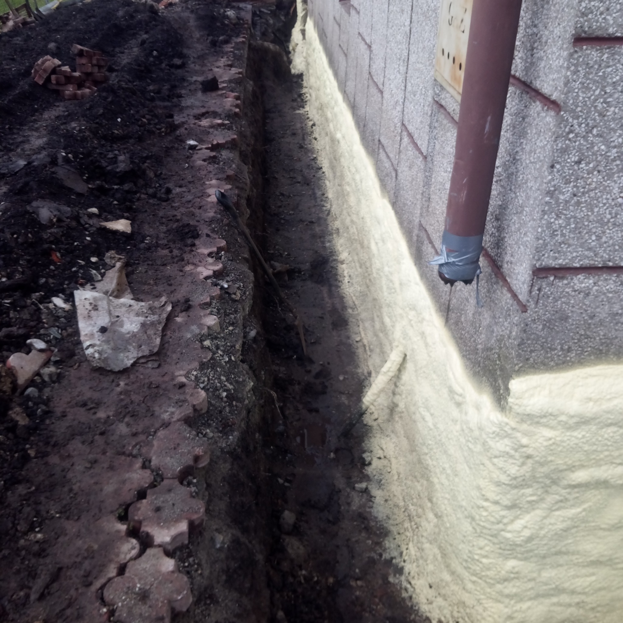 Wykonana izolacja pionowa fundamentu pianą PUR, widoczny wykop, cegły i rura spustowa z taśmą.