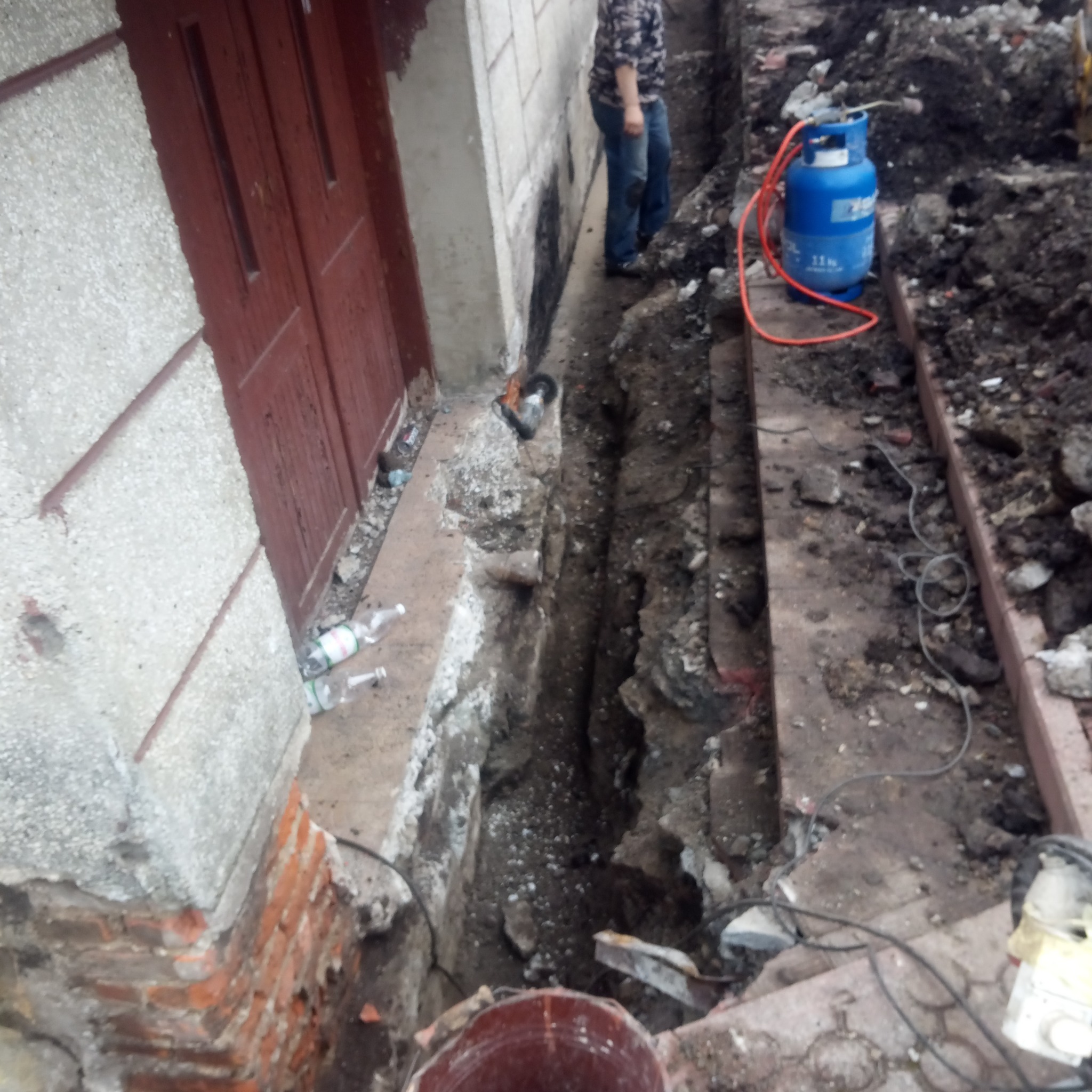 Wąski wykop wzdłuż ściany budynku z widoczną butlą gazową, narzędziami i osobą w tle, sugerujący prace ziemne i instalacyjne.