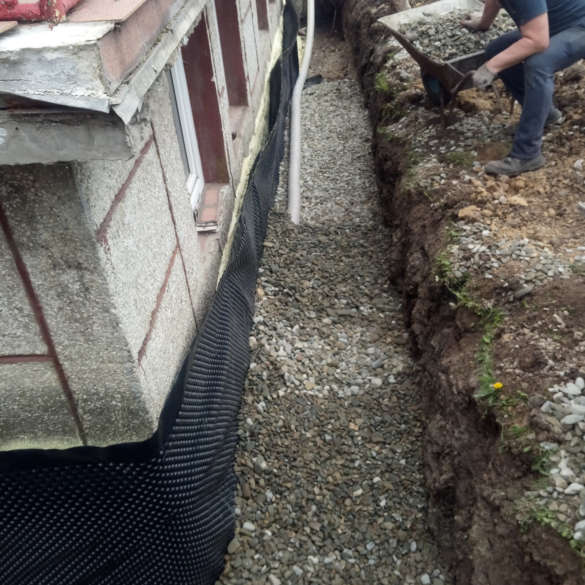 Wykop wzdłuż fundamentu budynku, zabezpieczony czarną folią izolacyjną, wypełniony żwirem; robotnik z taczką dosypuje żwiru.