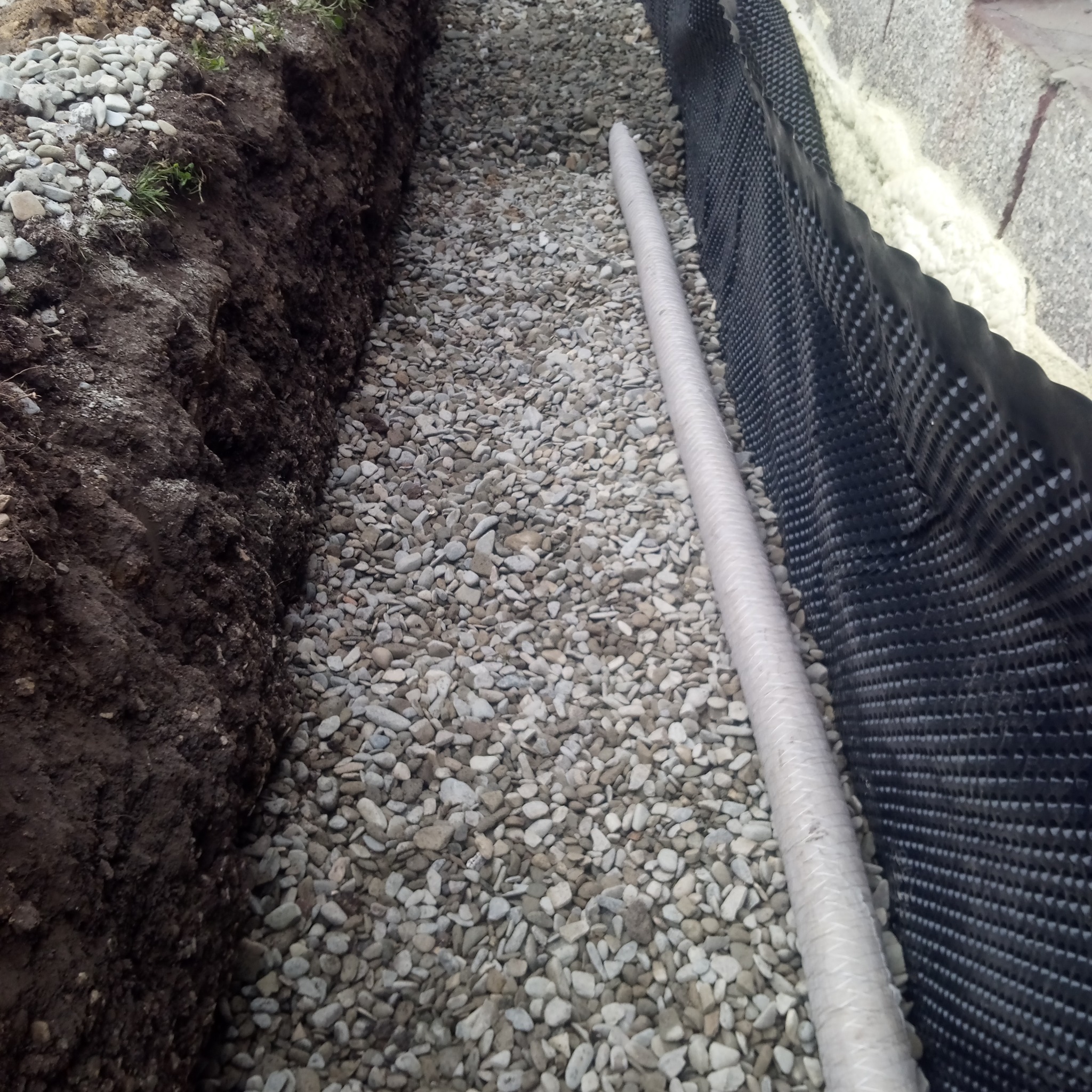 Wykop z rurą drenażową obsypany żwirem, zabezpieczony folią kubełkową przy fundamencie.